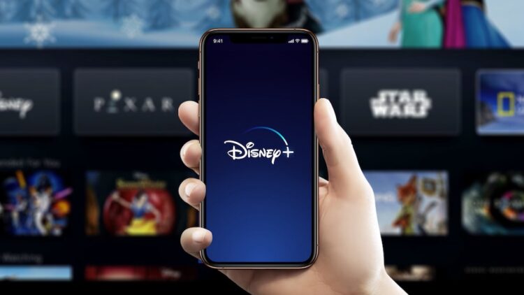 Brazil / November 18, 2019: Man holds an iPhone displaying the Disneyplus.com app and in the background Disney+ screen on TV. Disney+ is an online video streaming subscription service.